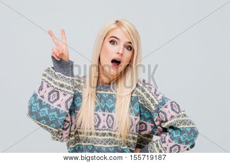Excited young blonde woman in sweater showing victory sign isolated on the gray background