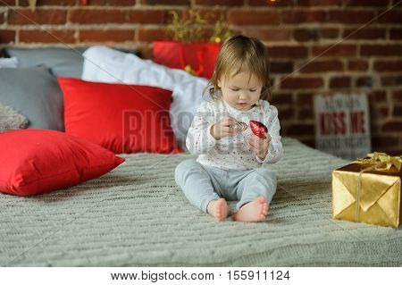 Christmas. The little girl sits on a bed. Nearby the Christmas gift in gold wrapping paper lies. The child with interest examines Christmas-tree decorations. Merry Christmas and a Happy New Year.