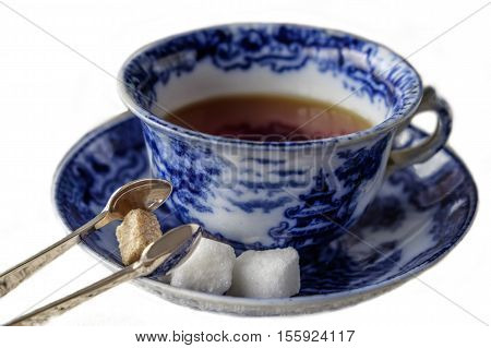 Antique blue and white china cup and saucer of black English breakfast tea with silver tongs for white and brown sugar cubes. Isolated on white background with copy space.