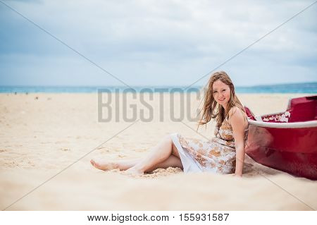 A Young Girl Sits On The Beach Leaning Against A Boat