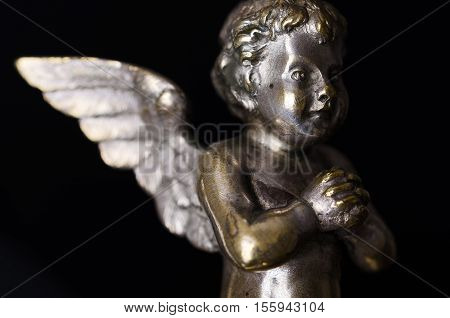 Praying brass angel side view over black. A winged putto made of brass, covered with silver, as part of a candelabra from the nineteenth century and a symbol for religious passion. Macro object photo.