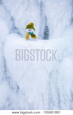 Snowman the Santa's assistant is standing in green hat wrapped with the scarf on nice textured snow on frosty winter morning.