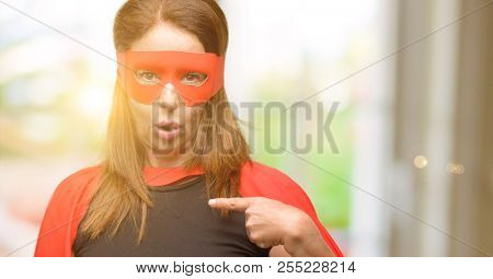 Middle age super hero woman wearing red mask and cape happy and surprised cheering expressing wow gesture, pointing with finger