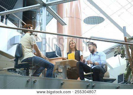 Group Of Young People In Casual Wear Sitting At The Office Desk And Discussing Something While Looki