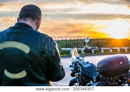 Biker In A Leather Jacket Standing Next To The Motorbike On The Background At Sunset