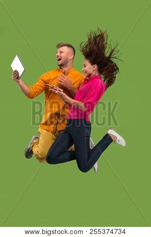 Jump Of Young Couple Over Green Studio Background Using Laptop Or Tablet Gadget While Jumping.