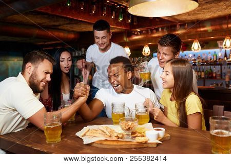 Two Handsome Male Friends Having Arm Wrestling Challenge At Bar. Friends Standing Around And Emotion