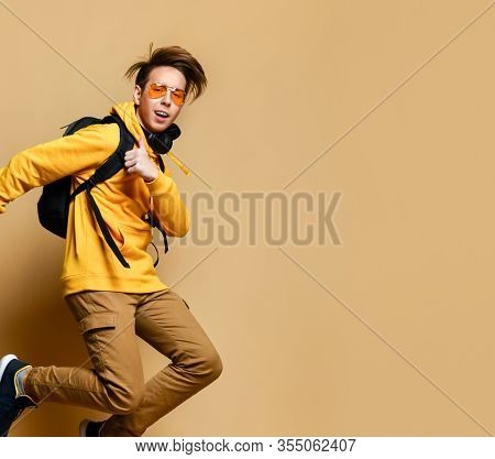 Young Teenager Boy In Comfortable Clothes, Sneakers, Sunglasses And A Backpack Jumping Over A Yellow