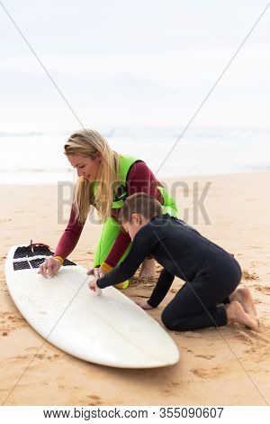 Mother And Son Waxing Surfboard On Beach. Happy Mother And Little Son In Wetsuits Sitting On Sand An