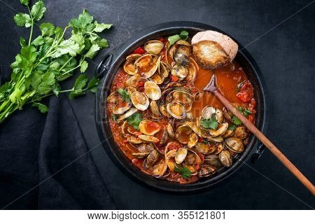 Cooked Japanese carpet shell with paprika and baguette in spicy tomato garlic sauce as top view in a modern design pot 
