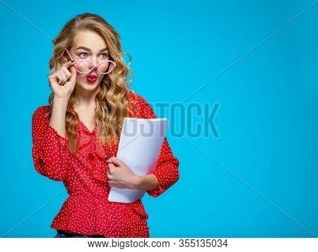 Surprised businesswoman in a casual clothes holds papers, at studio. Shocked woman secretary in a red shirt over blue background holding documents.  Female student in a pink glasses holding documents 
