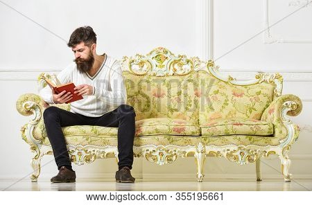 Self Education Concept. Man With Beard And Mustache Sits On Baroque Style Sofa, Holds Book, White Wa