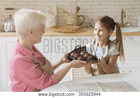 Home Cooked Meals. Smiling Girl And Her Granny Holding Tray With Fresh Chocolate Muffins At Home