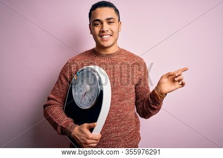 Young brazilian man doing diet to lose weigth holding scale over isolated pink background very happy pointing with hand and finger to the side