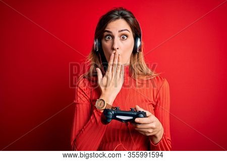 Young beautiful brunette gamer woman playing video game using joystic and headphones cover mouth with hand shocked with shame for mistake, expression of fear, scared in silence, secret concept