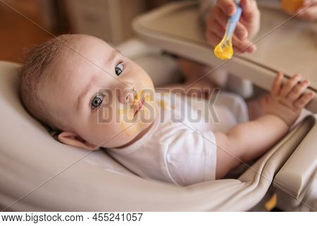 Mother Feeding Her Baby Boy Using Spoon, Introducing First Solid Food Meal, Baby Sitting In High Cha