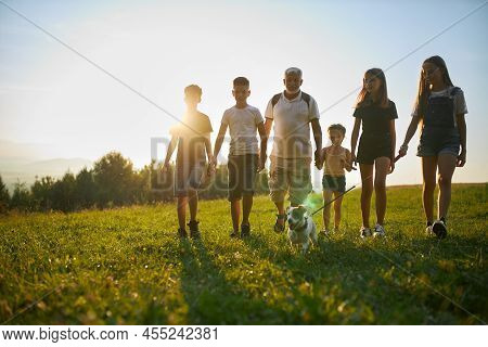 Front View Of Children With Old Man And Dog Walking In Mountains. Male With Gray Hair And Beard With