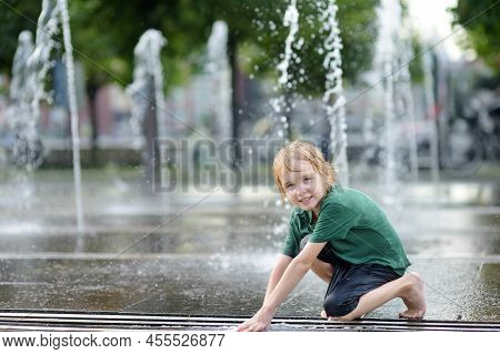 Little Boy Plays In The Square Between The Water Jets In The Ciry Fountain At Sunny Summer Day. Acti