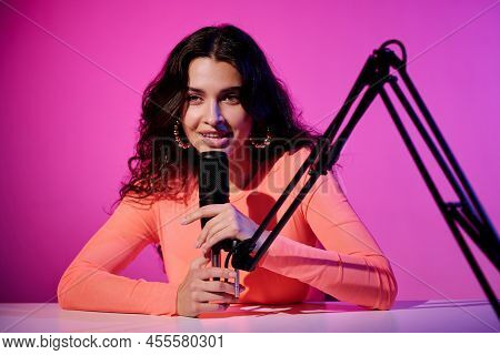 Beautiful Young Female Influencer Sitting At Desk In Studio In Pink Neon Light Whispering In Microph