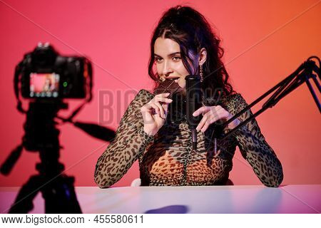 Attractive Young Caucasian Woman Sitting In Studio In Pink And Orange Neon Light Recording Chocolate
