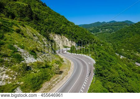 Asphalt Road Is Meandering Between Blue Sea And Green Mountains. Aerial View Of Car Driving Along Th