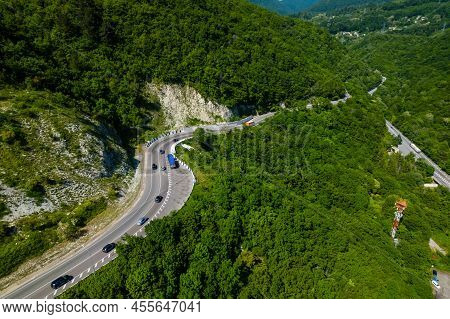 Asphalt Road Is Meandering Between Blue Sea And Green Mountains. Aerial View Of Car Driving Along Th