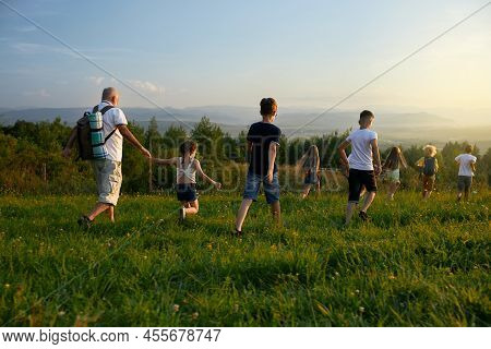 Back View Of Kids With Old Man Leading Each Other Forward In Hills. Male With Gray Hair Wearing Ruck
