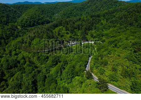Asphalt Road Is Meandering Between Blue Sea And Green Mountains. Aerial View Of Car Driving Along Th