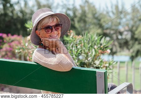 Portrait Of Beautiful Senior Woman Wearing Cap And Glasses Sitting Outdoor On A Bench Of Public Park