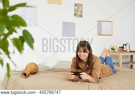 Happy Attractive Young Woman With Brown Hair Lying On Bed With Djambe And Using Smartphone While Wat