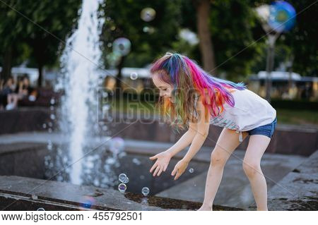 Cute Little Caucasian Girl With Colorful Dyed Hair Catching Soap Bubbles By Splashing Fountain In Ci