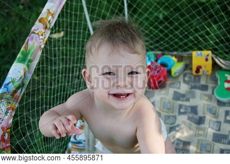 Cute Little Baby Who Can't Walk Yet Is In Play Pen. Naked Baby Standing In An Play Pen, Looking Up A