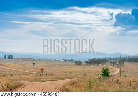 Country Unsealed Gravel Dirt Road Surrounded By Farms And Fields In Australia. Mountains On The Hori