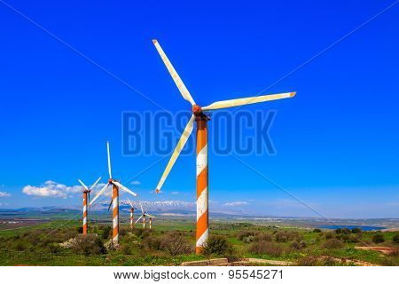 Israel. Several huge modern windmills. Seen in the distance the snow-covered Hermon. Flowering Golan Heights on a sunny day