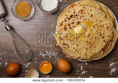 Flapjack on brown wooden table with flour, eggs and whisk