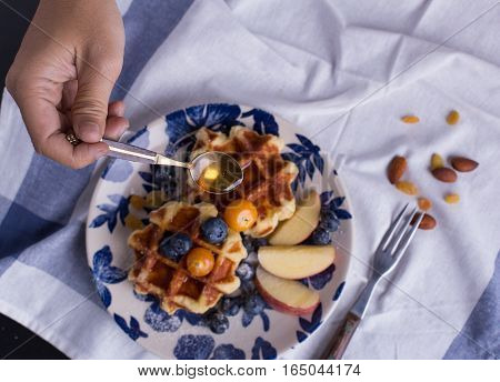 Blueberry apple waffle with honey set on a dish with a chef's hand holding spoon for a jar of honey ready to serve.