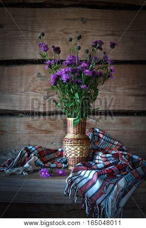 Beautiful purple wild flowers cornflowers in a straw vase on a wooden bench in an old farmhouse