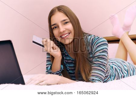 Young beautiful smiling girl makes online shopping using laptop computer lying on bed. Pink background.