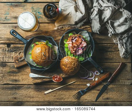 Homemade beef burgers with crispy bacon and fresh vegetables in small cast iron pans and glass of wheat beer on rustic board over old wooden background, top view, horizontal composition