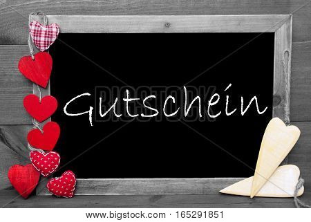 Chalkboard With German Text Gutschein Means Voucher. Red Hearts. Wooden Background With Vintage, Rustic Or Retro Style. Black And White Image With Colored Hot Spots.