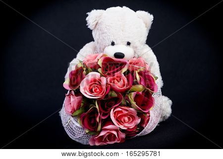 Selective focus at bouquet of beautiful artificial red roses in the arms of blurry white bear doll on black background concept love for valentine day or spacial anniversary