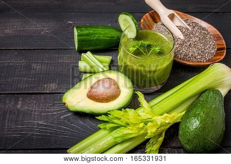 Healthy green juice smoothie surrounded by avocado, cucumber,  celery and chia seeds on black wooden background, top view