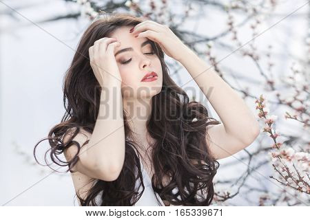 Sensual young girl standing in white dress at blossoming tree in the garden against white background. Beauty of woman and nature consept. Portrait of beautiful model with curly brown hair sensual posing with closed eyes.