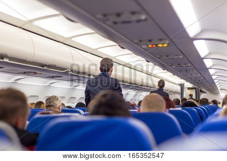 Interior of airplane with passengers on seats and stewardess walking the aisle.