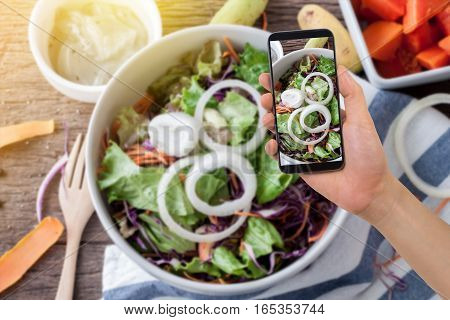 human hand hold smartphone tablet cell phone on blurry kitchen table with vegetable salad and dressing concept of diet food take a photo vegetable salad on screen cell phone.