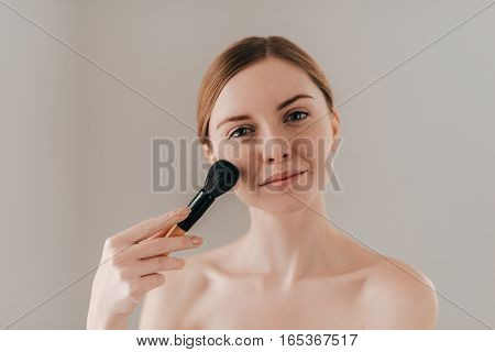 Make-up for natural look. Beautiful young woman holding make-up brush and looking at camera while standing against background
