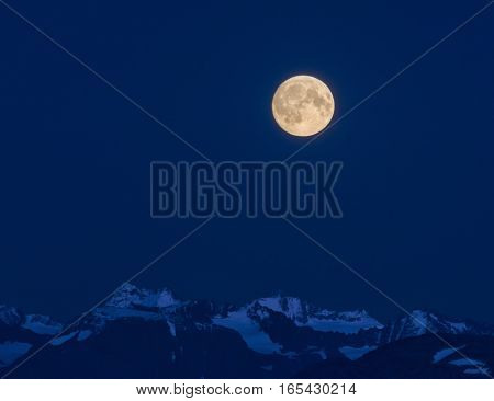 A full moon shines bright white over the blue shrouded Kenai Mountains.