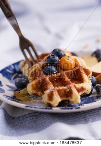 Closeup of a fork cutting through a blueberry waffle decorated with fresh blueberry in a nice dish.