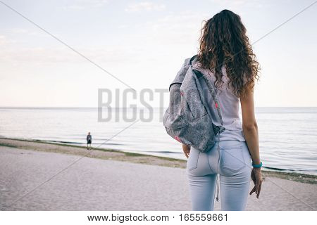 Young Sporty Woman With Curly Hair, Wearing Jeans And A Backpack Standing On The Beach And Looking A