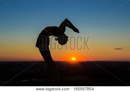 Silhouette of sporty woman practicing yoga in the park at sunset - drop back, wheel pose. Sunset light, sun lens flares, golden hour. Freedom, health and yoga concept